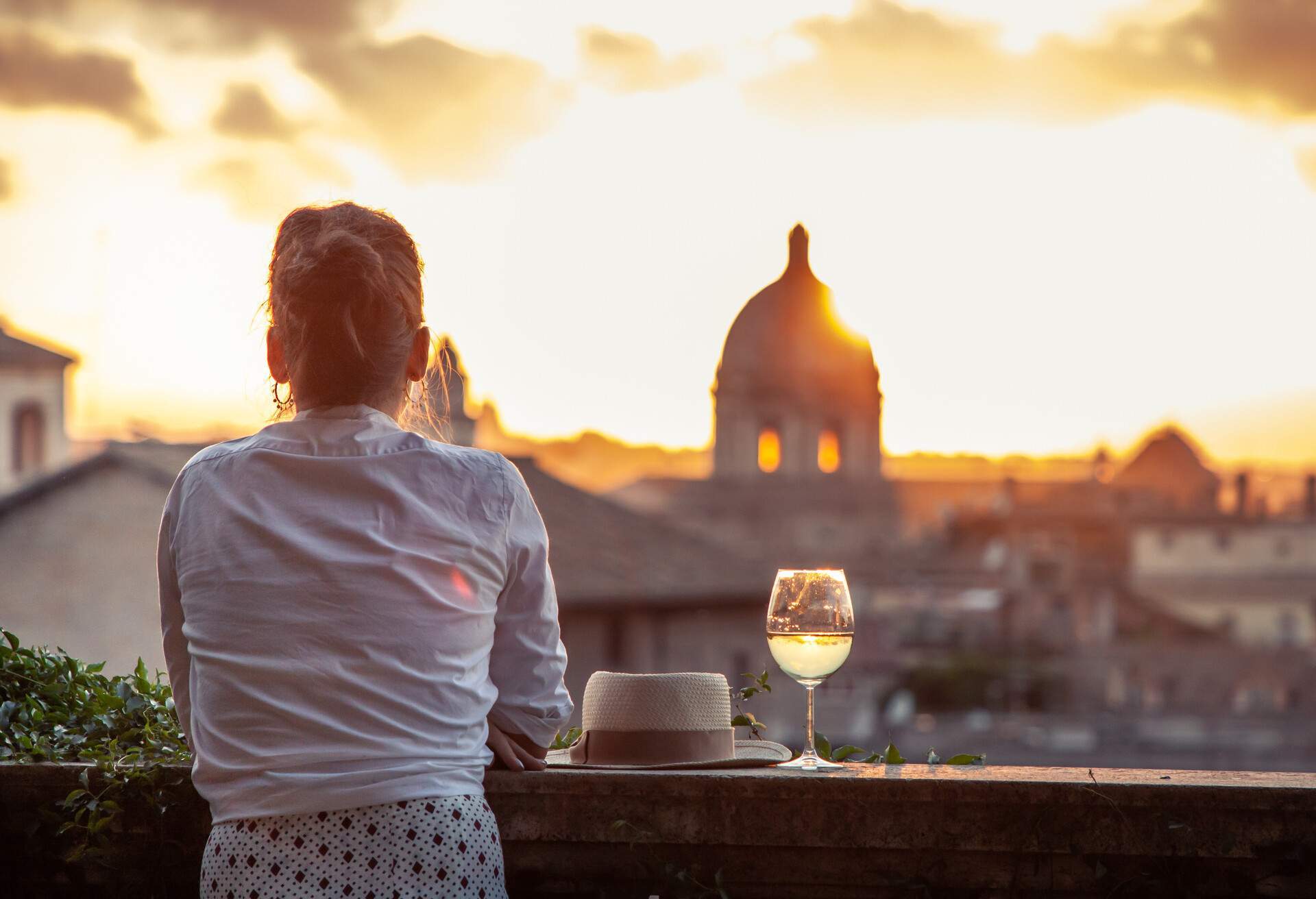 A woman leaning on a balcony railing next to a hat and a glass of wine as the sun sets over the city.