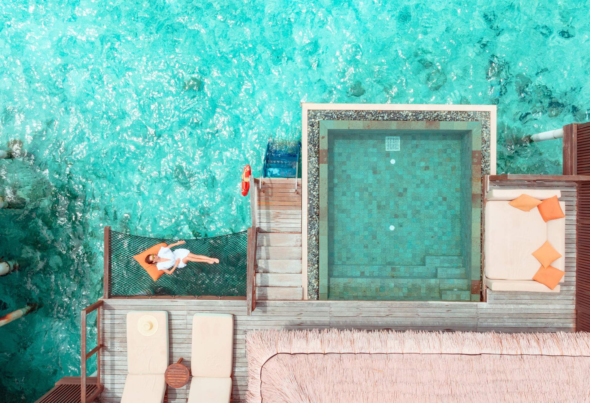 An overwater villa with a woman relaxing on a net bed beside a square pool with a couch and sunbeds.