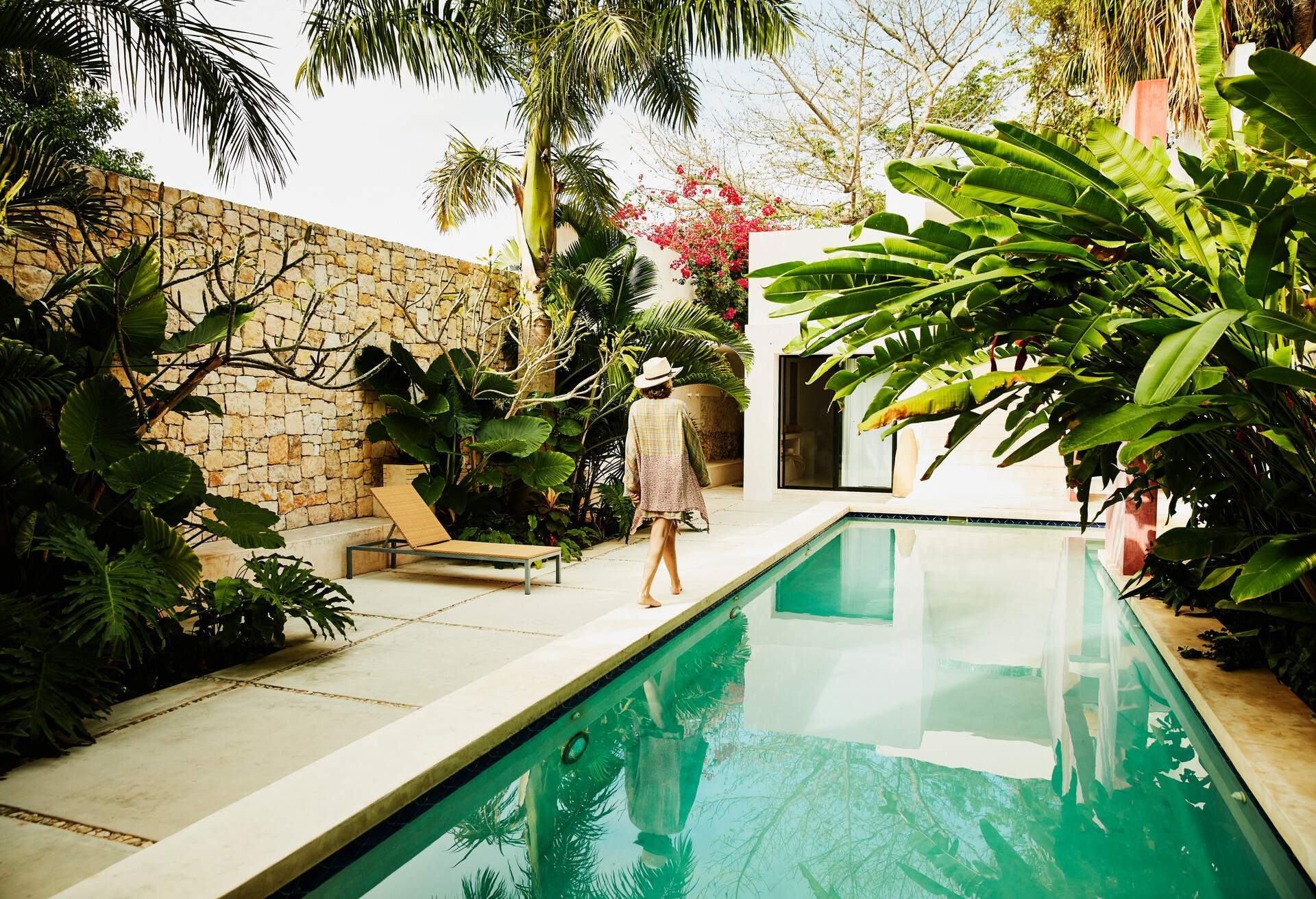 A woman strolls leisurely along the edge of a tranquil pool, with a wall framing the area and adding a touch of privacy and seclusion to the picturesque setting.