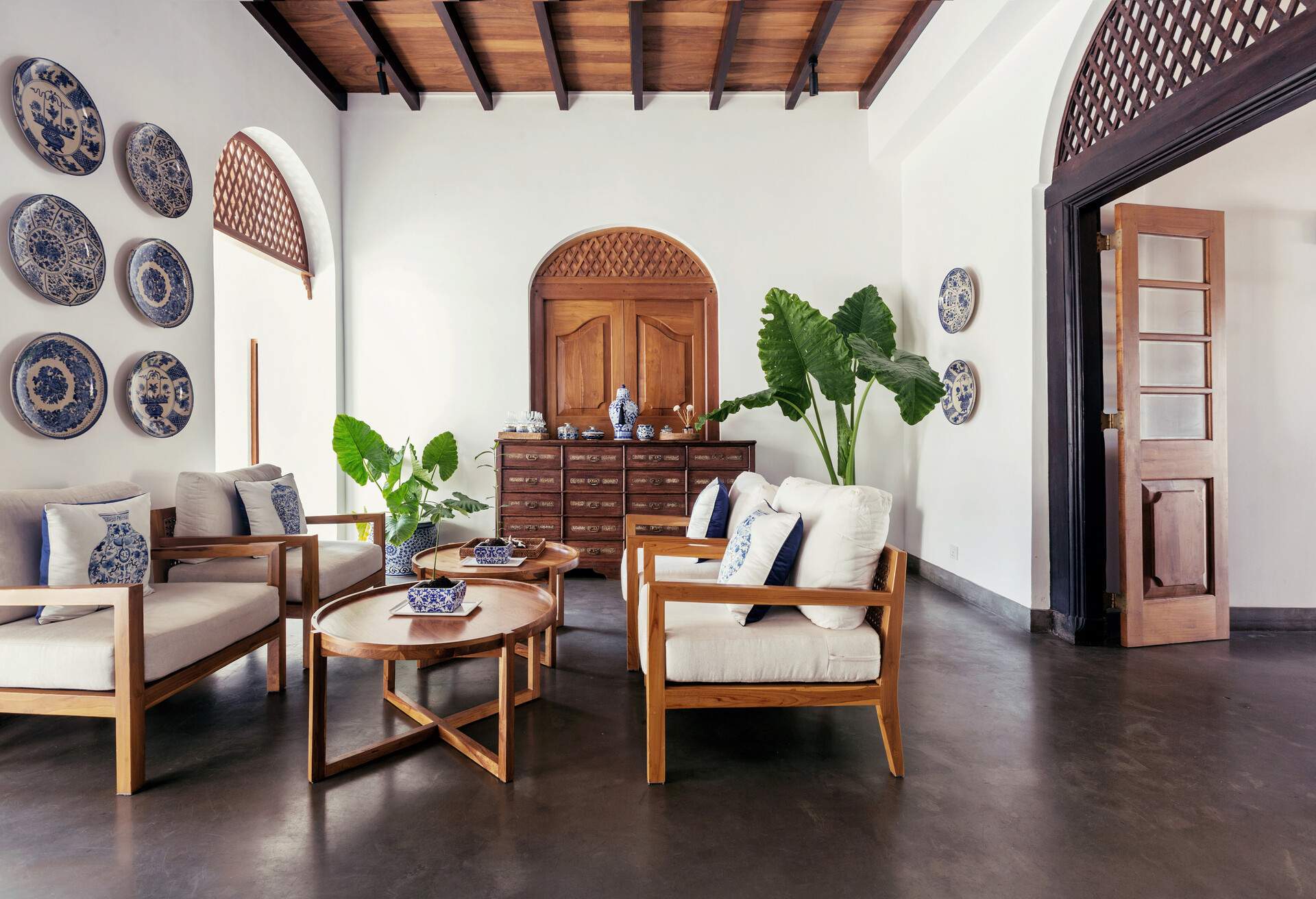 A light and bright living area with seats, wooden coffee tables, potted plants, and decorative wall plates.
