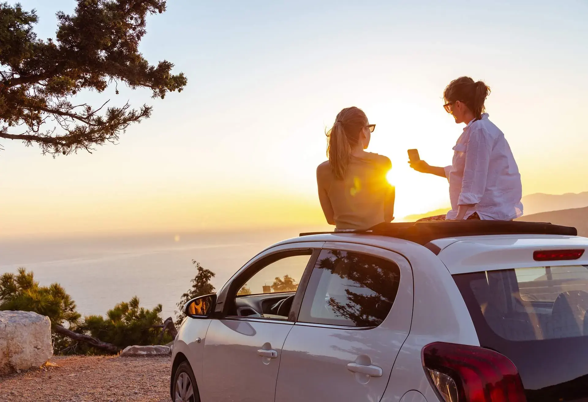 Mujer sentada sobre el techo de un auto blanco tomando una selfie al atardecer en un paisaje natural con árboles.