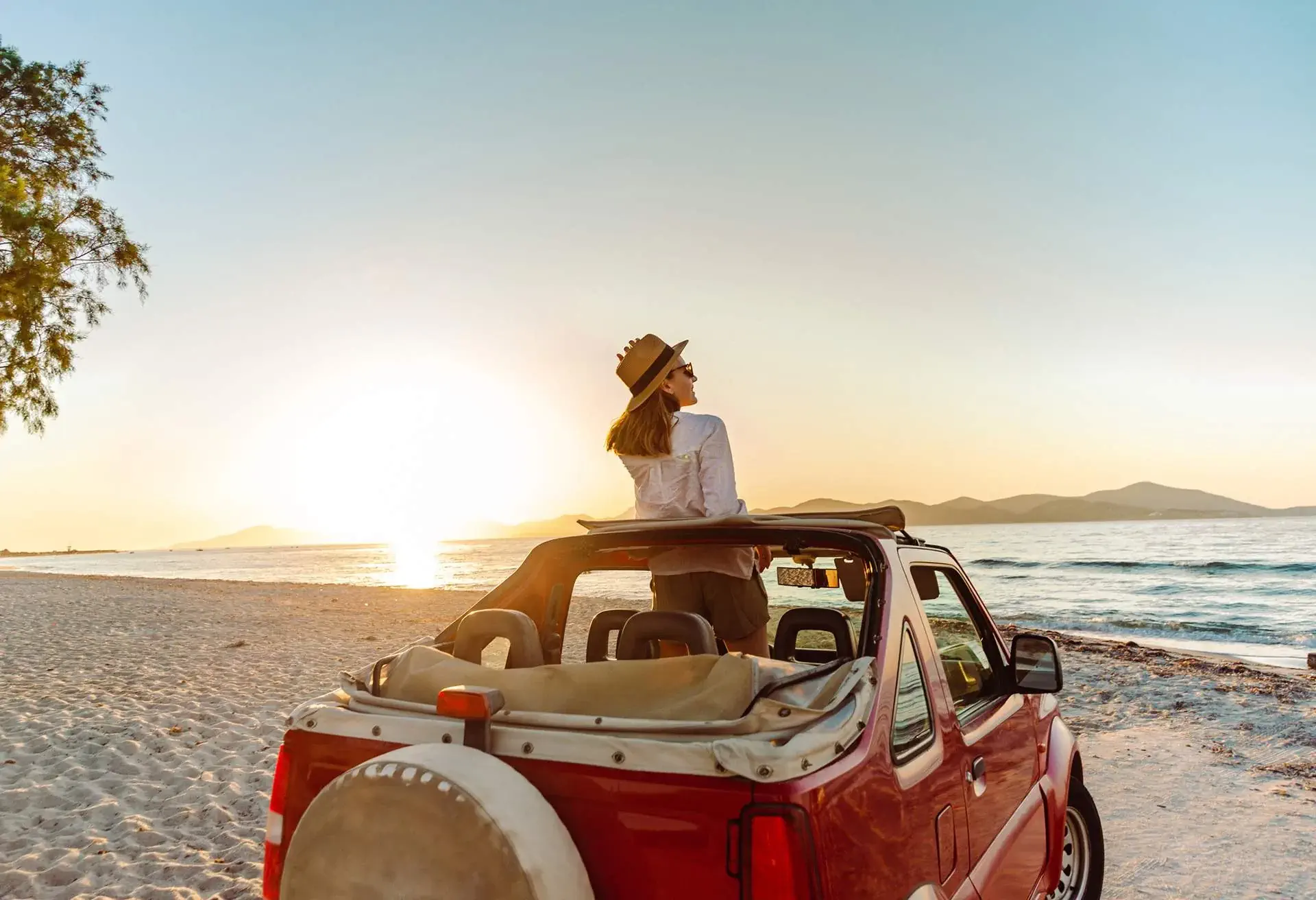 Mujer de pie en un auto rojo descapotable estacionado frente al mar, mirando el atardecer en una playa tranquila.