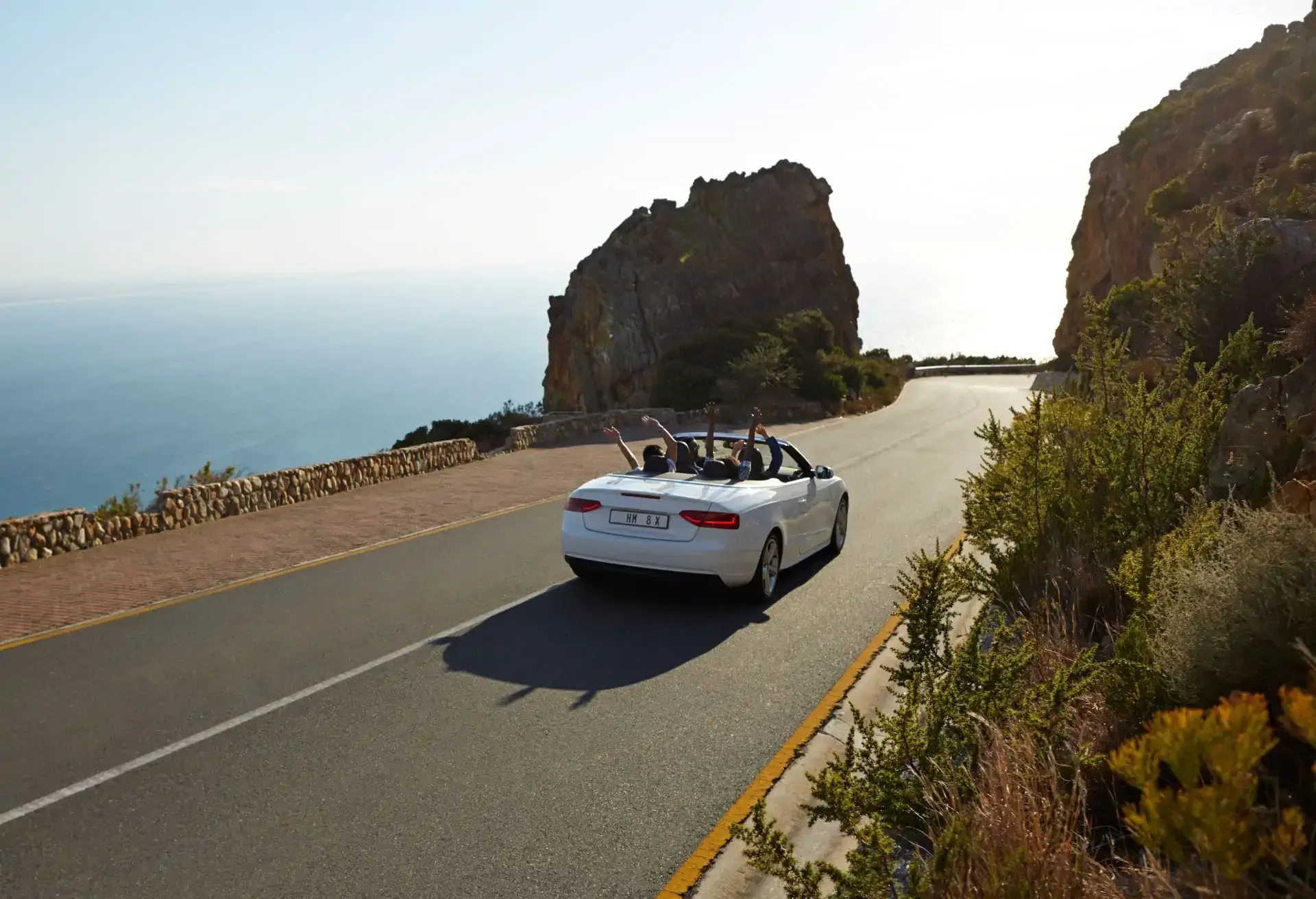 Auto convertible blanco manejando por una carretera costera con acantilados y vista al mar al atardecer.