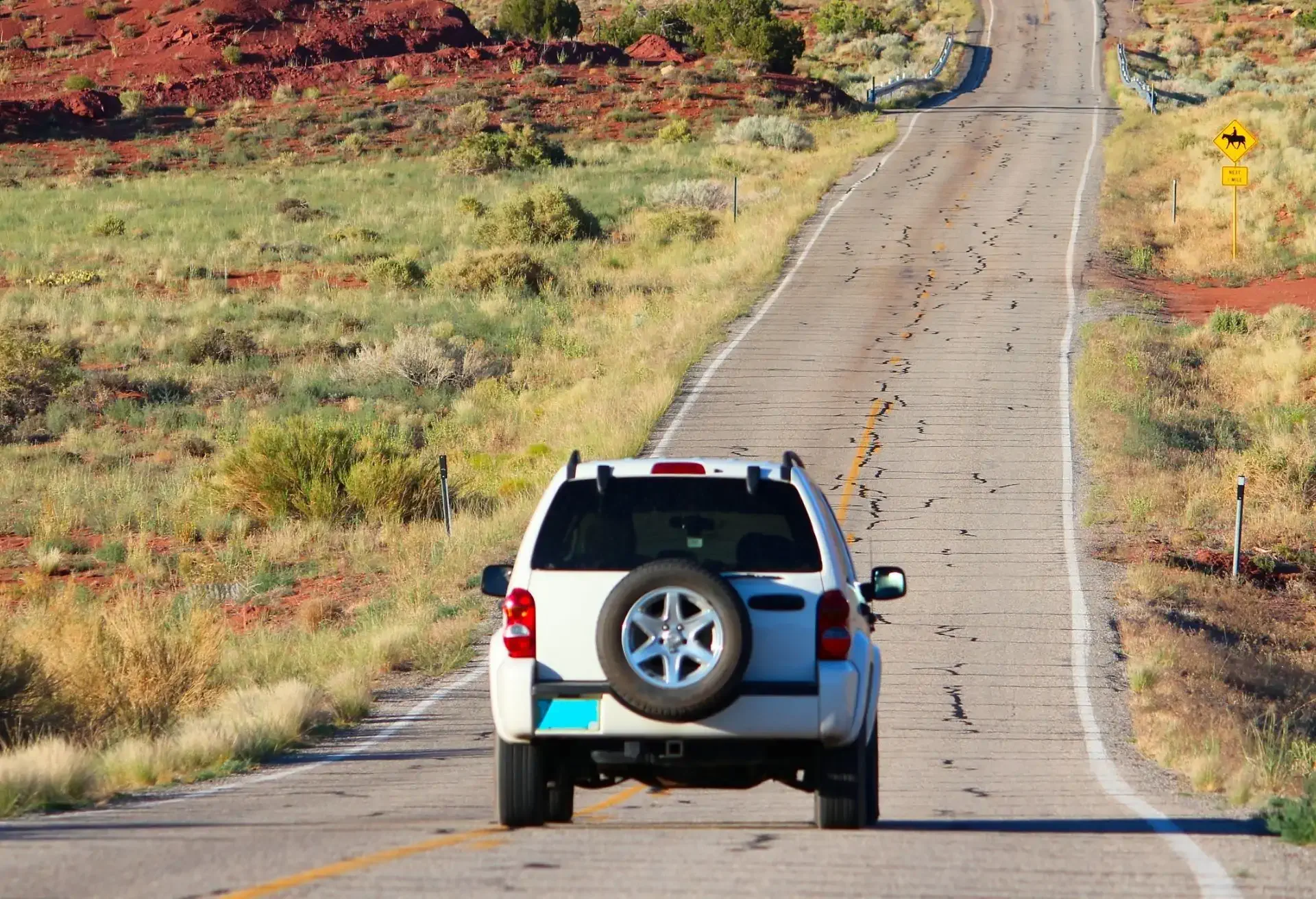 Vehículo SUV blanco avanzando por una carretera solitaria en un paisaje semidesértico con vegetación baja y montañas rojas al fondo.