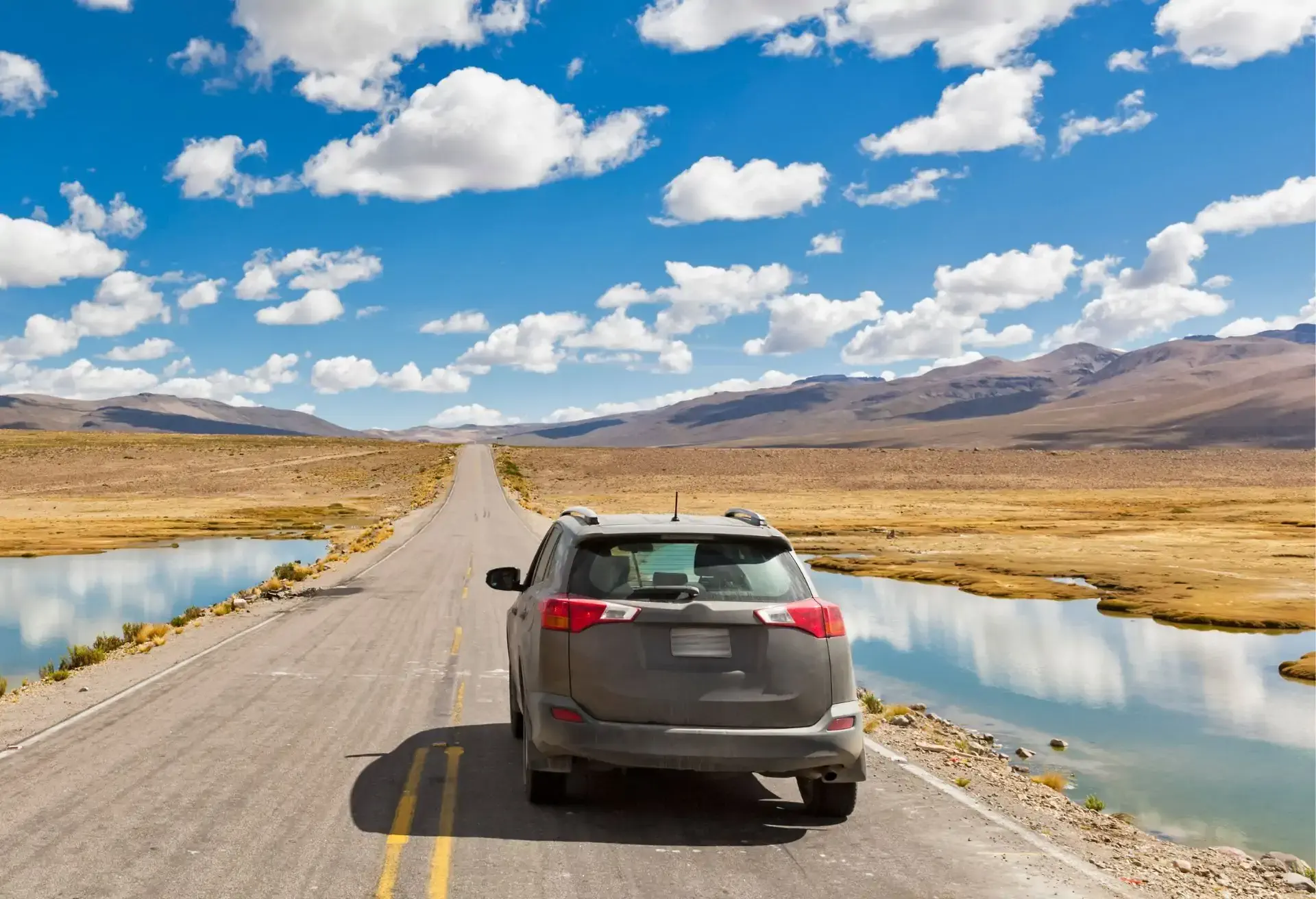 Automóvil gris transitando por una carretera desierta junto a un lago cristalino en un paisaje montañoso y abierto.