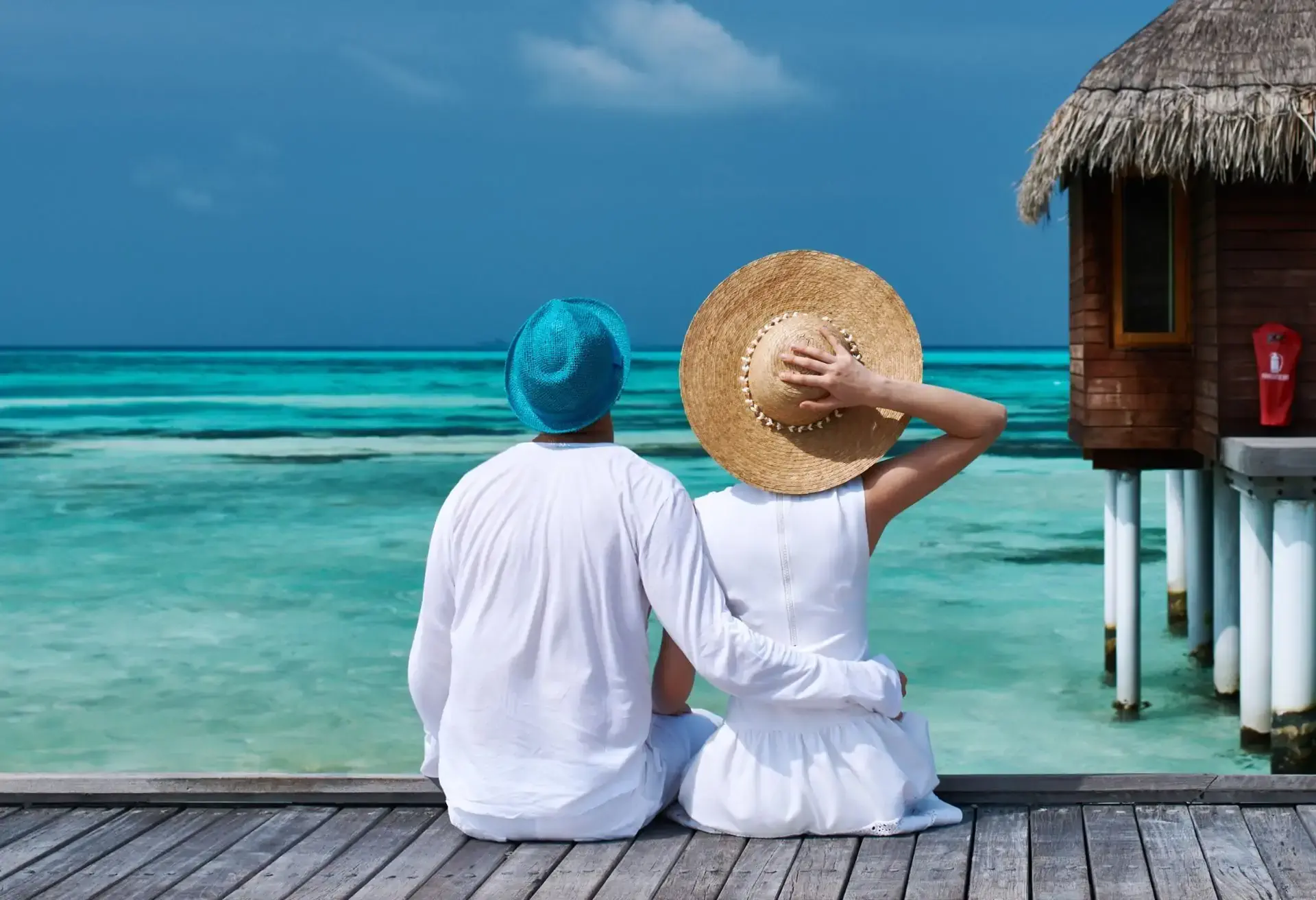 Pareja sentada al borde de un muelle de madera mirando el mar turquesa, con sombreros de verano en un destino exótico.