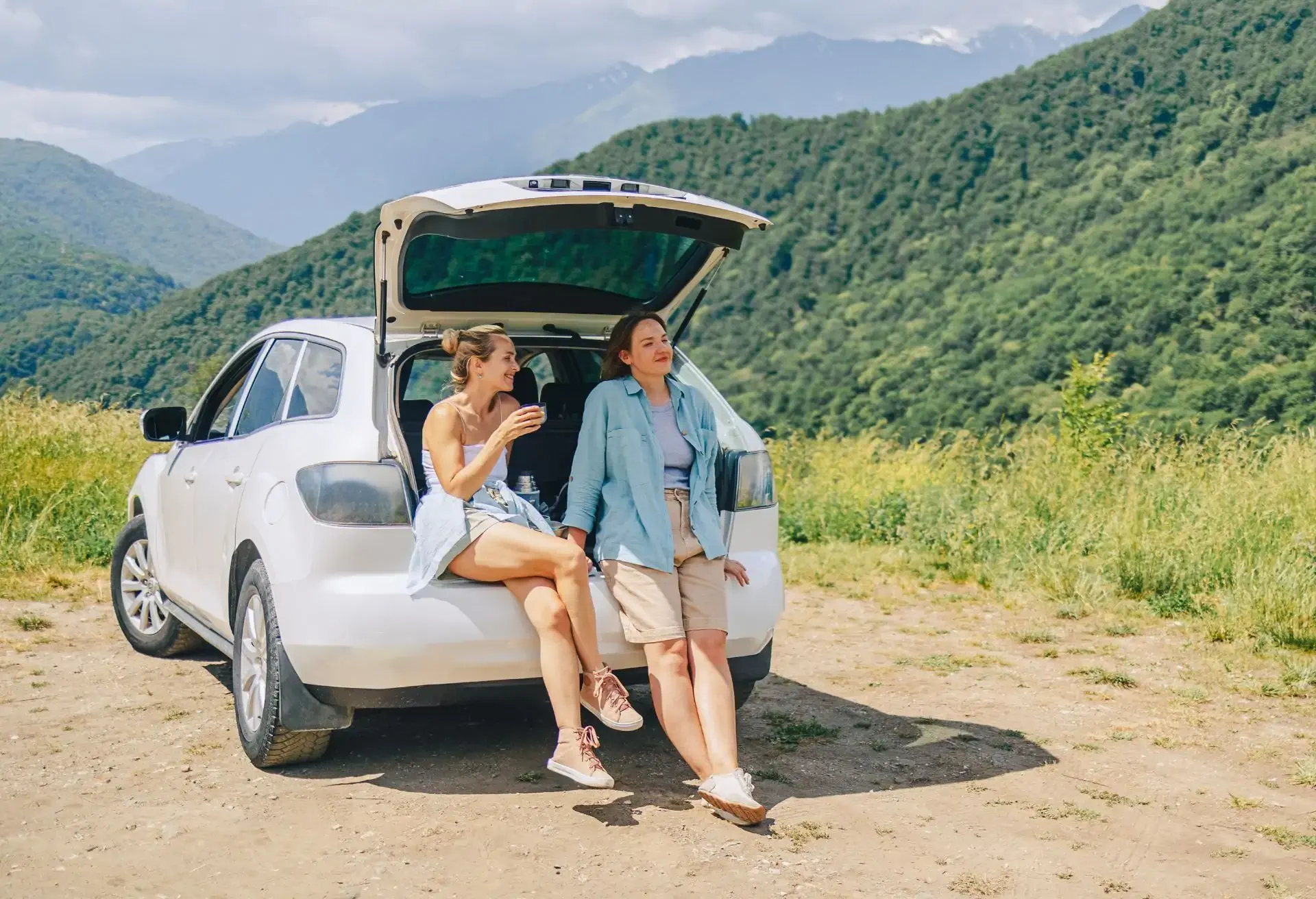 Dos mujeres conversando junto a un auto blanco con la cajuela abierta en una zona boscosa durante un viaje por carretera.