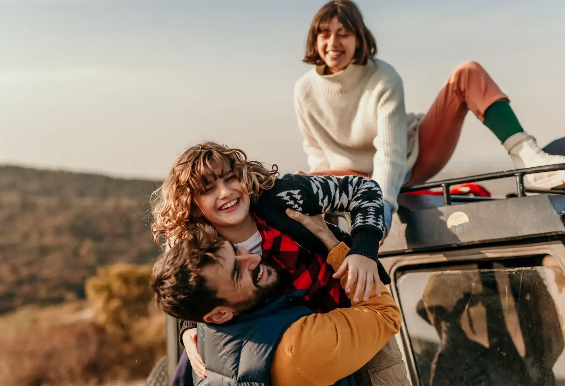Padres e hija riendo y jugando sobre una manta junto a un auto en un campo abierto durante un paseo familiar.