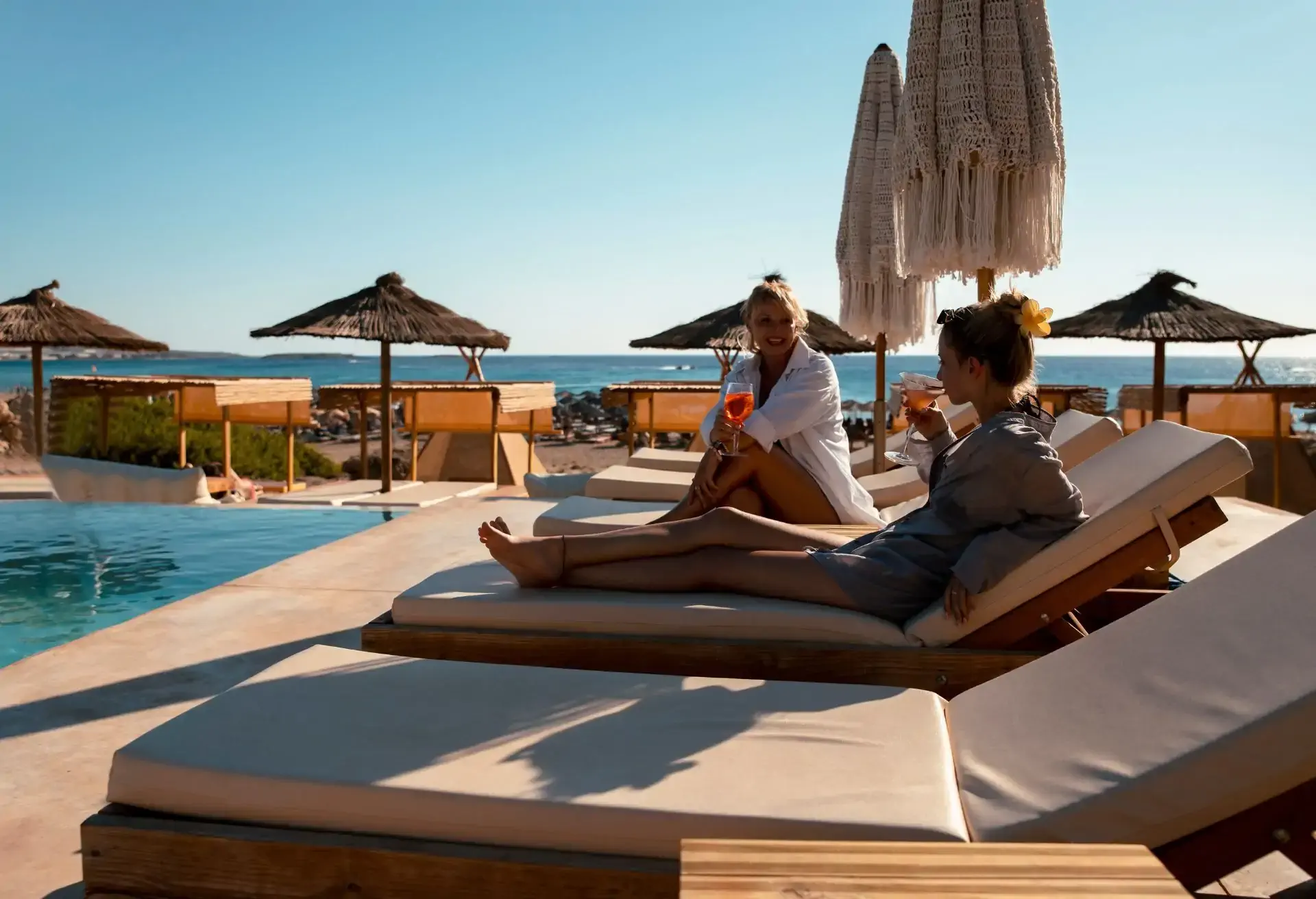 Personas relajándose en camastros junto al mar, bajo sombrillas de palapa, disfrutando de un día soleado frente a la playa.