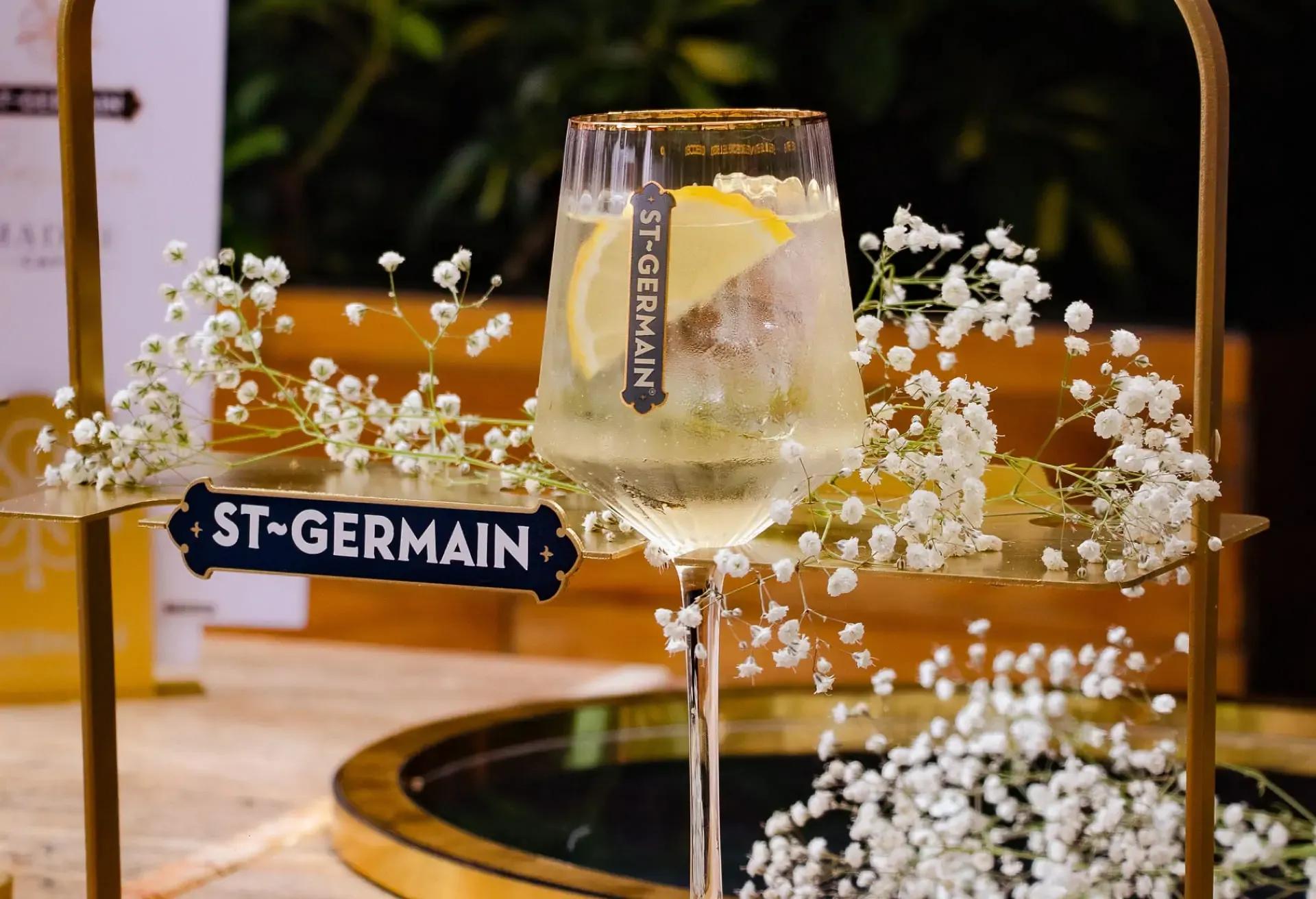 Copa de St-Germain Spritz con rodaja de limón decorada con flores blancas sobre una bandeja dorada.