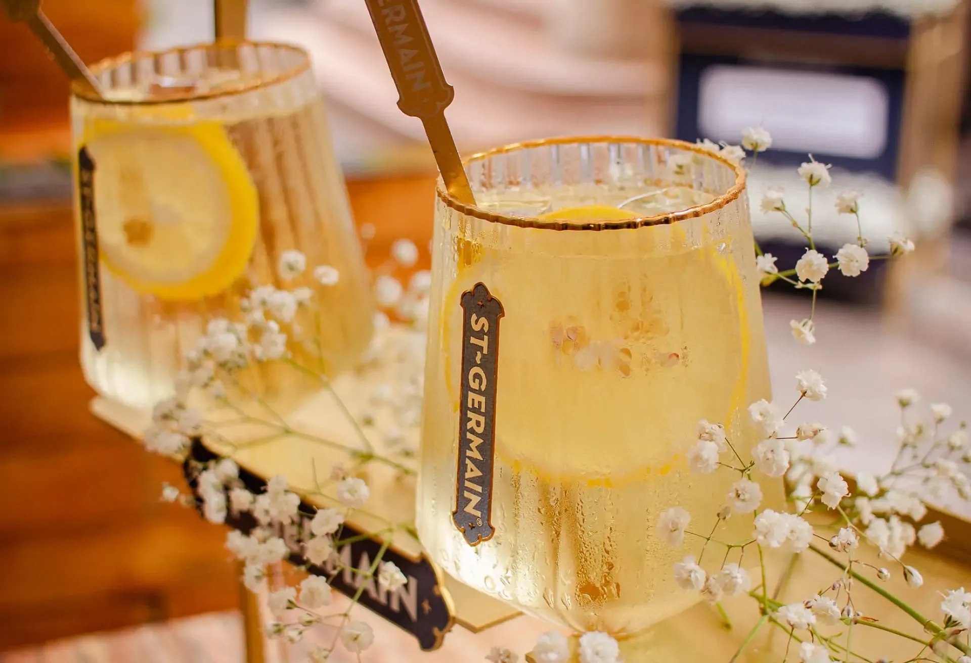 Copas de St-Germain Spritz con rodajas de limón y flores blancas decorativas sobre una bandeja dorada.