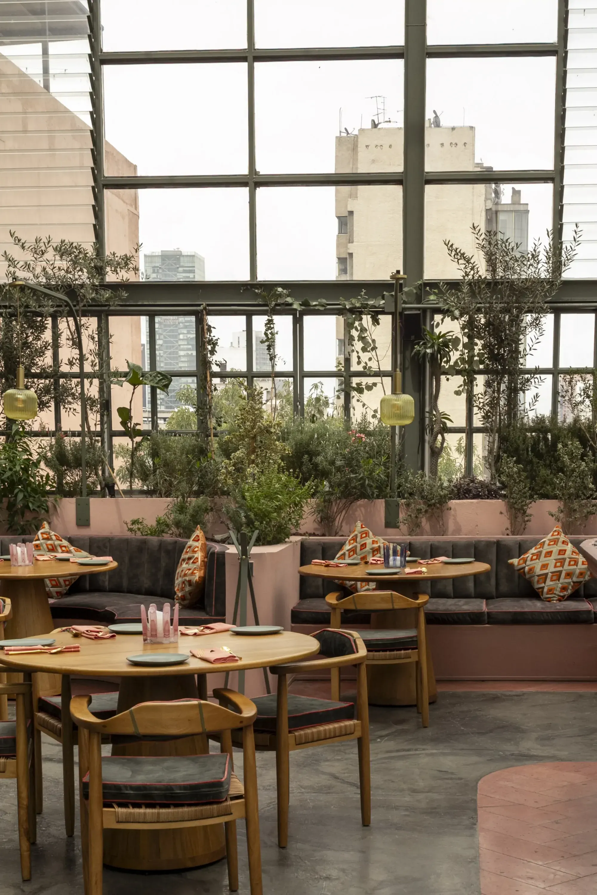 Terraza interior con mesas de madera, vegetación y ventanales con vista urbana.