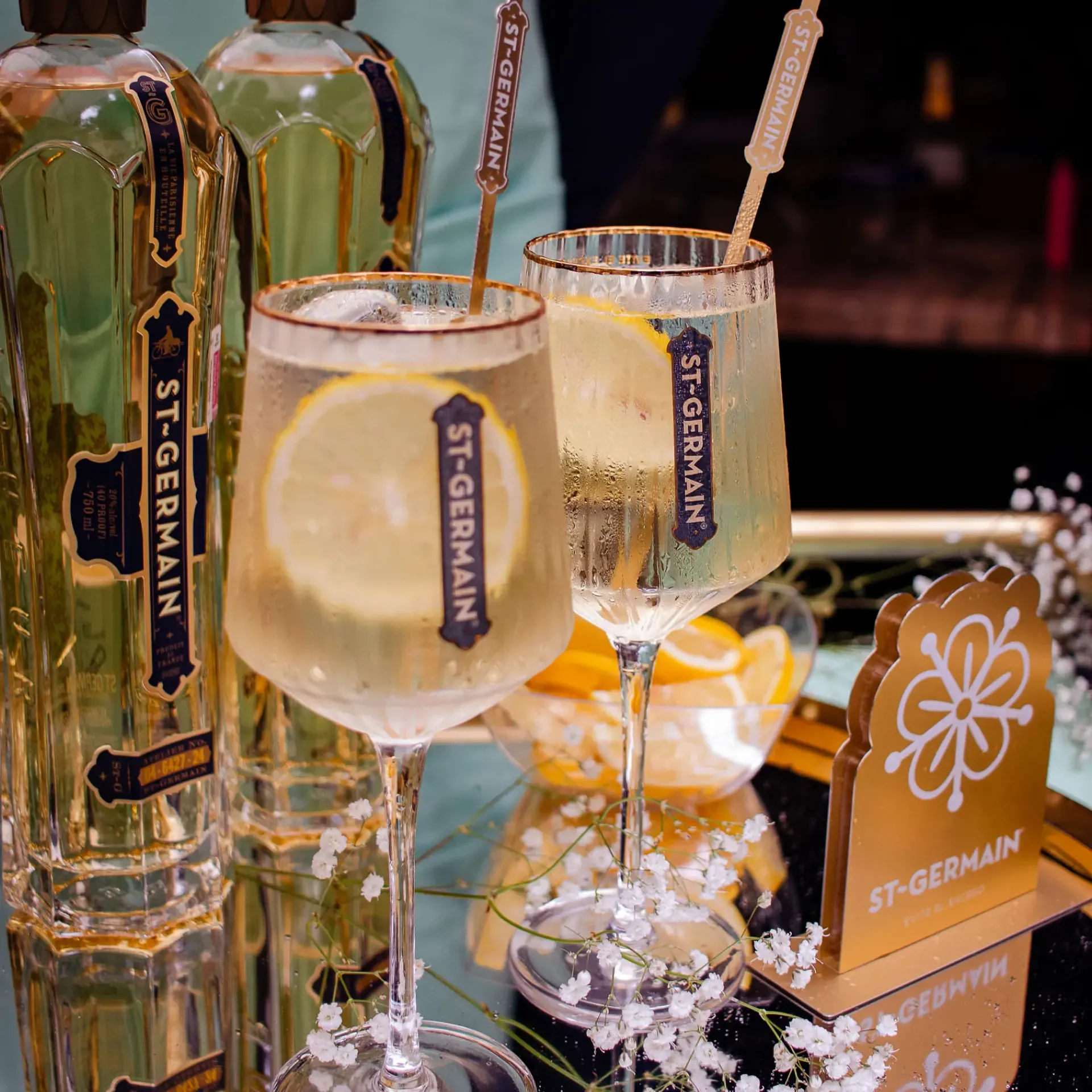Copas de St-Germain Spritz con rodajas de limón junto a botellas del licor y decoración floral sobre una mesa elegante.