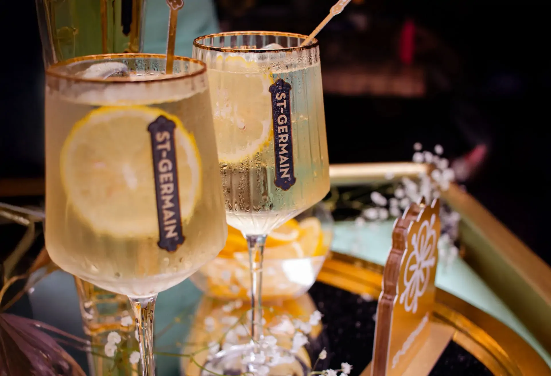 Copas de St-Germain Spritz con rodajas de limón sobre una bandeja dorada decorada con flores blancas.