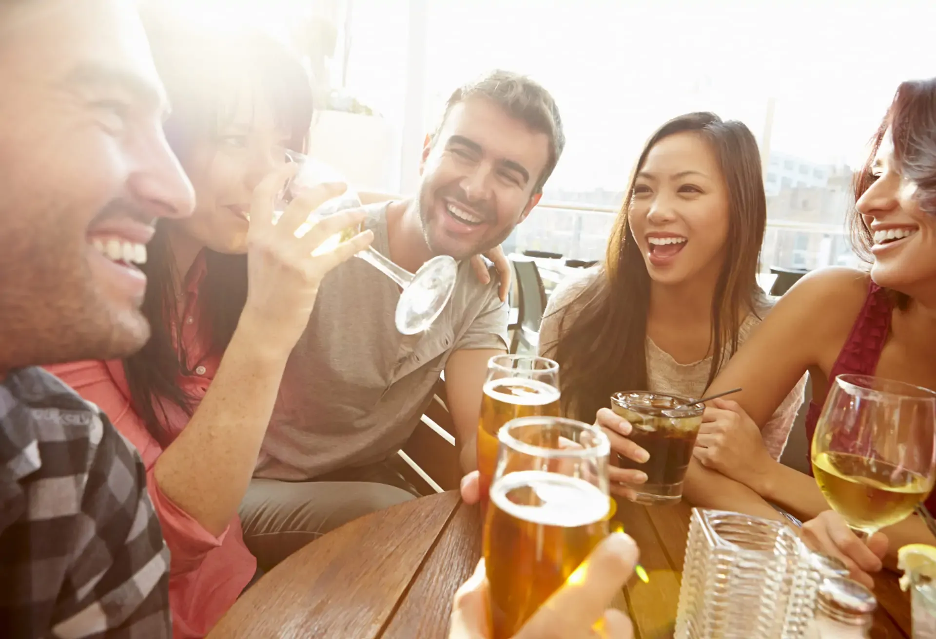 Grupo de amigos riendo y brindando con bebidas en una terraza iluminada por el sol.