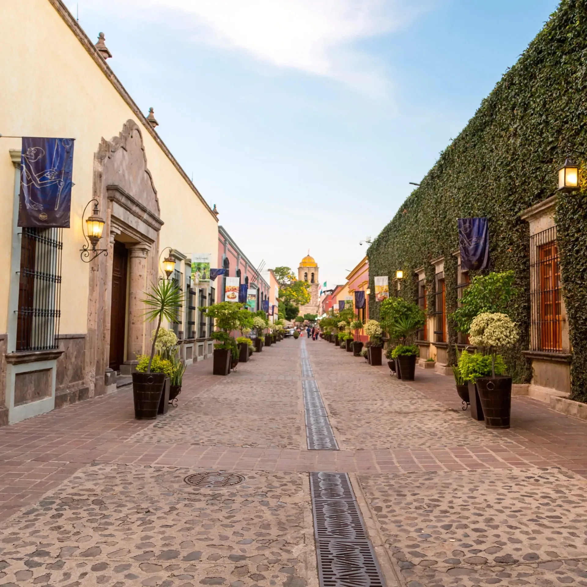 Calle empedrada con fachadas coloniales, plantas y una cúpula amarilla al fondo en un día soleado.
