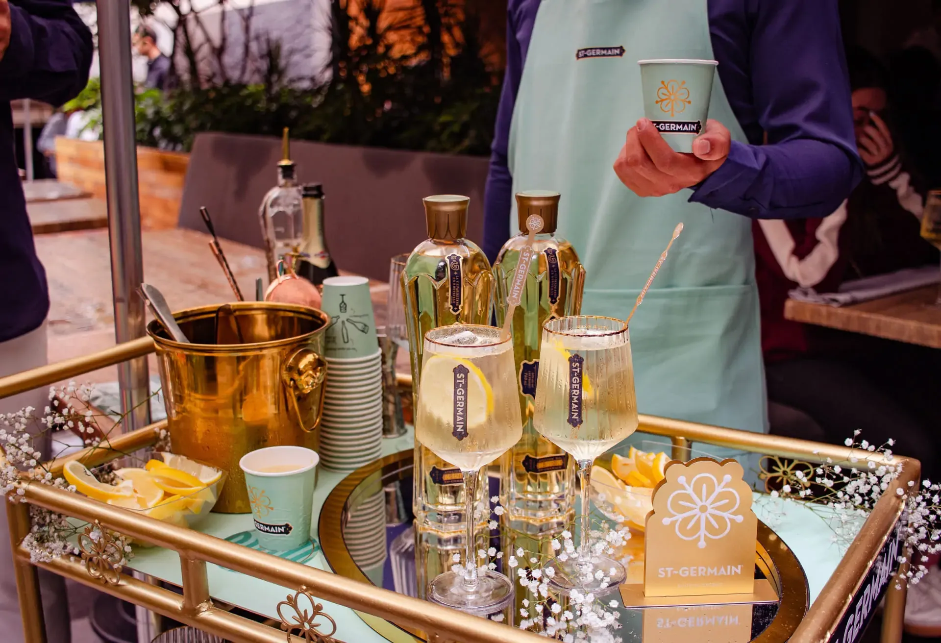 Carrito con botellas, copas y elementos de St-Germain Spritz atendido por una persona con delantal color verde menta.