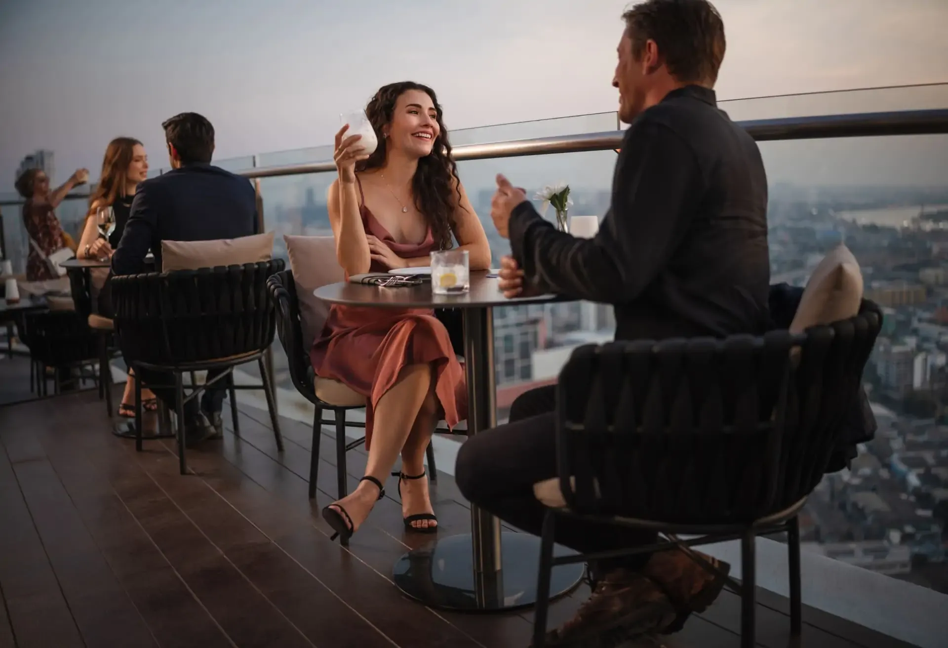 Personas conversando y disfrutando bebidas en una terraza elevada con vista panorámica al atardecer.