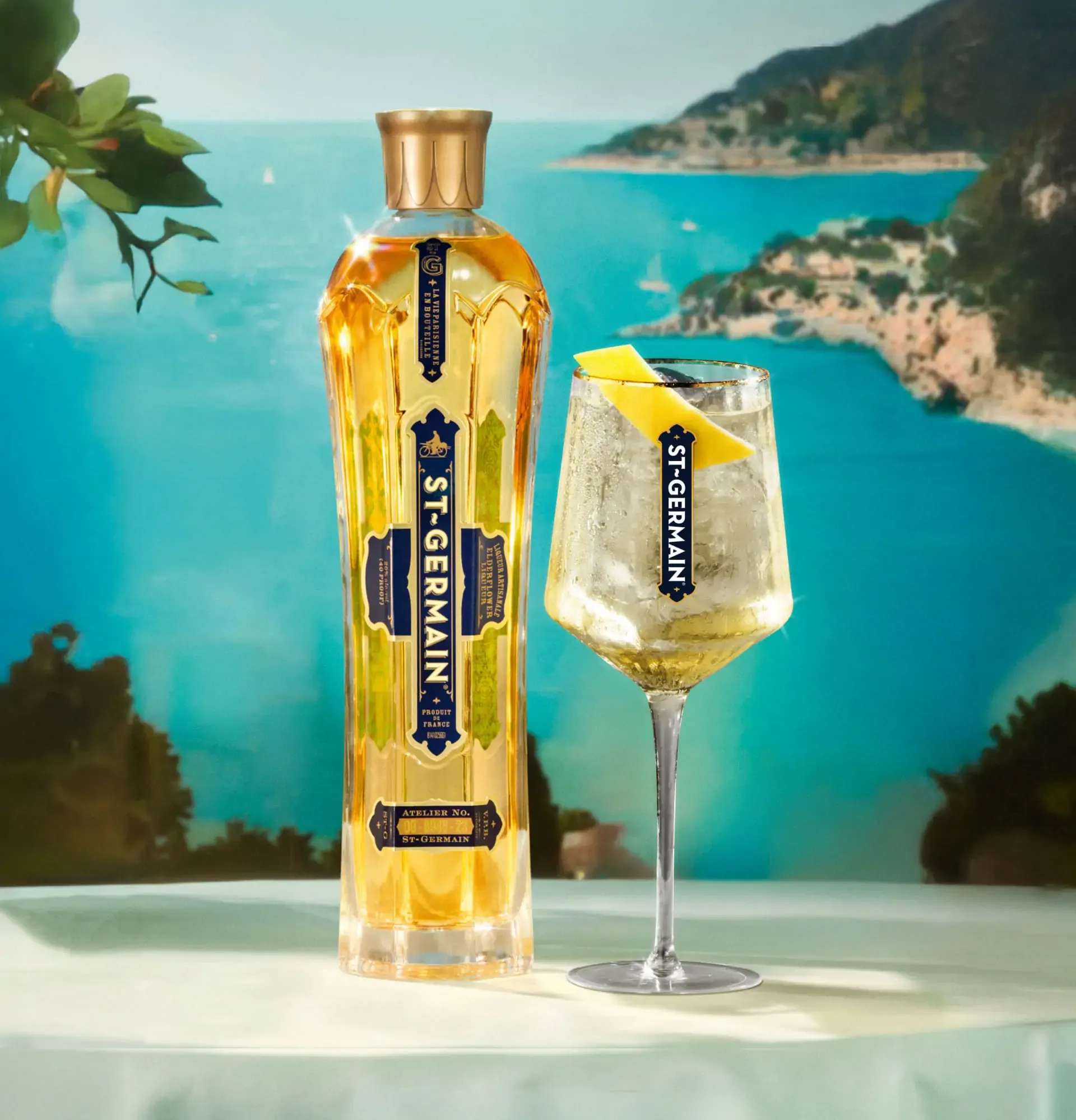 Botella alta y elegante de licor St-Germain de color dorado, con etiquetas azules y doradas, junto a una copa de tallo largo que contiene un cóctel de color pálido (posiblemente un spritz) adornado con una cáscara o rodaja de limón. El fondo es un paisaje costero brillante con mar turquesa y colinas verdes, evocando un ambiente veraniego y lujoso.